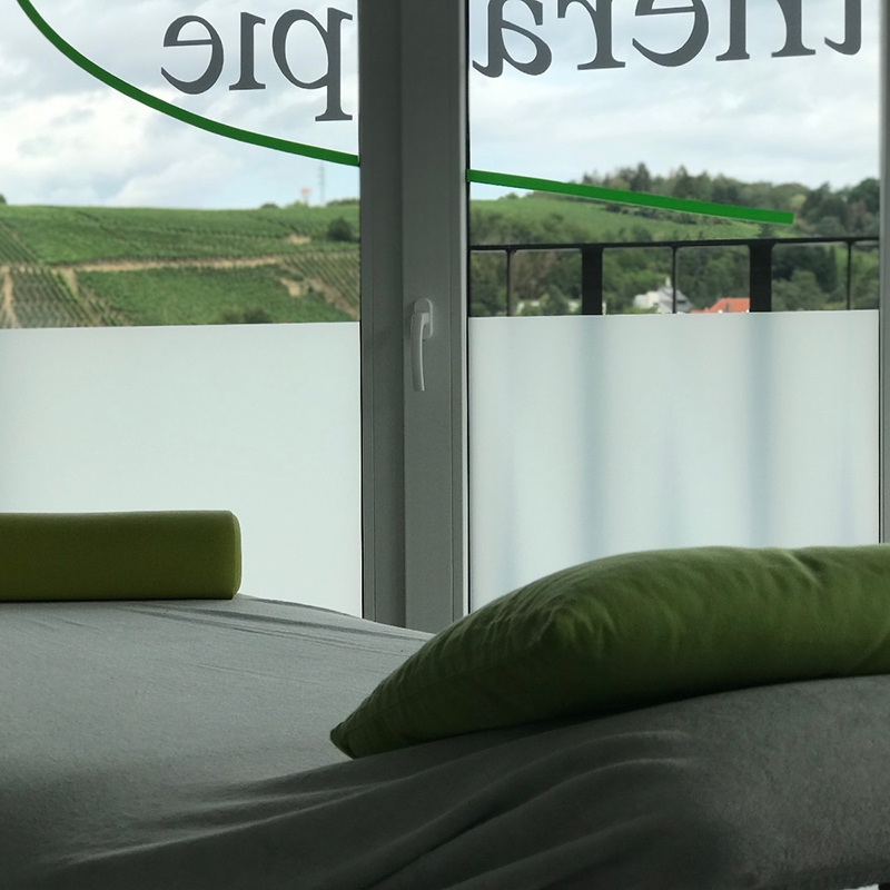 Massageliege mit grünen Kissen und Blick aus dem Fenster im Hintergrund.