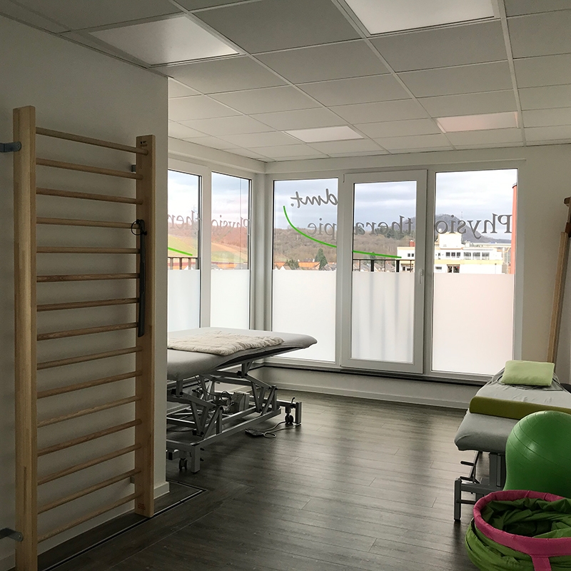 Behandlungsraum der dmt. Physiotherapie-Praxis mit Kletterleiter an der Wand und Massageliegen im Raum. Helle Fensterfront im Hintergrund und grünes Übungsgerät an der rechten Seite.