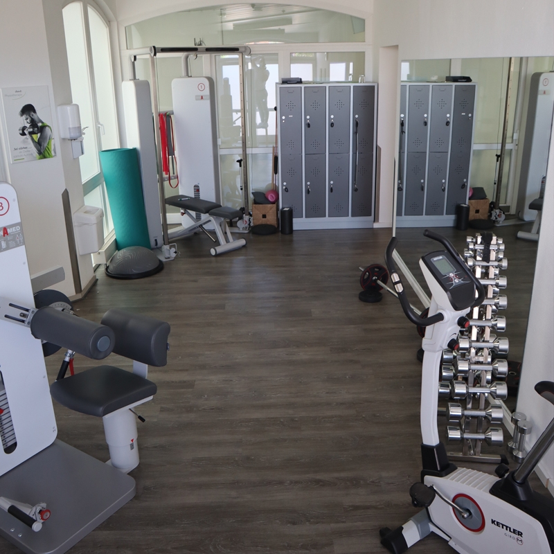 Trainingsraum mit Trainingsgeräten, ähnlich wie in einem Fitnessstudio. Links ist ein Spiegel zum Kurz- und Langhantel-Training. Daneben ein Turm mit Kurzhanteln und Gewichten am Boden. Abschließbare Spinte im Hintergrund.