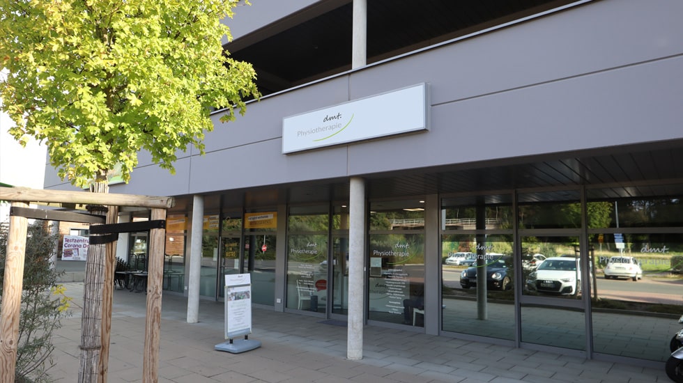 Außenansicht auf das Gebäude von dmt. Physiotherapie in Grafschaft. Schild mit Logo über dem Eingang in die Praxis. Hellgraue Fassade.
