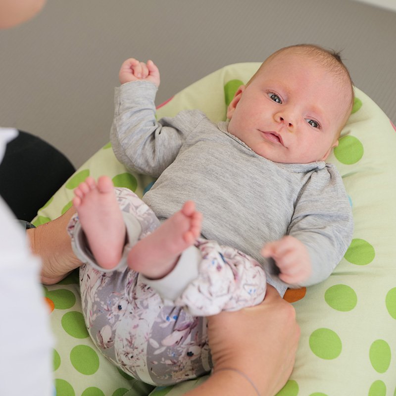 dmt. Kids. Physiotherapie an den Beinen, durchgeführt von einer Physiotherapeutin bei einem Kleinkind, dass in einem grünen Kissen liegt.