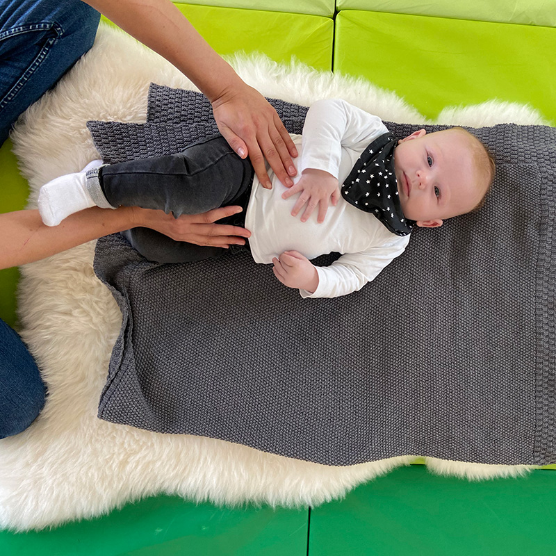 Hüft-und Beckenübung bei einem Kleinkind auf bequemer Unterlage durch die Mutter, angeleitet durch den Physiotherapeuten.