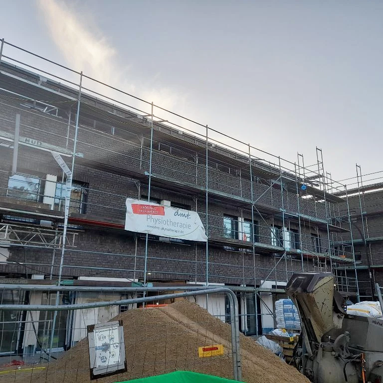 Das Bild zeigt das neue Praxisgebäude in Erftstadt. Es ist noch in ein Gerüst gekleidet, an dem ein Banner der dmt. Physiotherapie hängt.