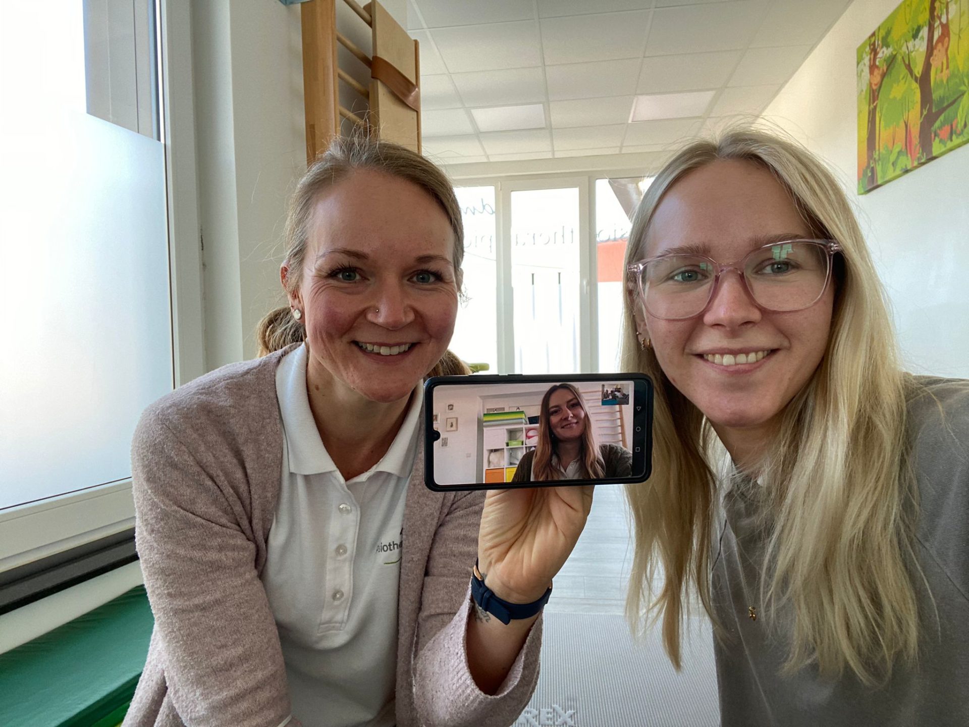 Links ist Corinna, rechts Anne. Corinna hält ein Handy in der Hand. Auf dem Display ist ein Bild von Jasmin zu sehen. Die drei machen einen Videocall.