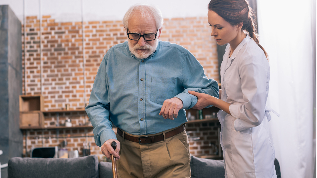 Die Physiotherapeutin stützt einen älteren Mann am Arm, der am Gehstock läuft. Er trägt ein blaues Hemd und eine braune Hose, Brille und hat graue Haare.