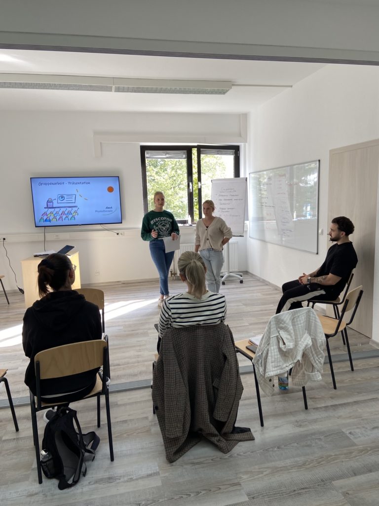 Mehrere Praktikanten sitzen im Stuhlkreis und schauen auf eine Leinwand. Die dmt. Physiotherapie Mitarbeiter halten einen Vortrag.
