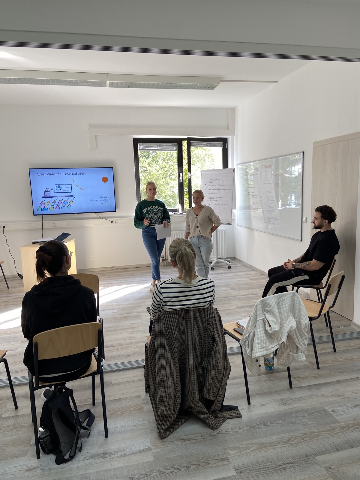 Mehrere Praktikanten sitzen im Stuhlkreis und schauen auf eine Leinwand. Die dmt. Physiotherapie Mitarbeiter halten einen Vortrag.