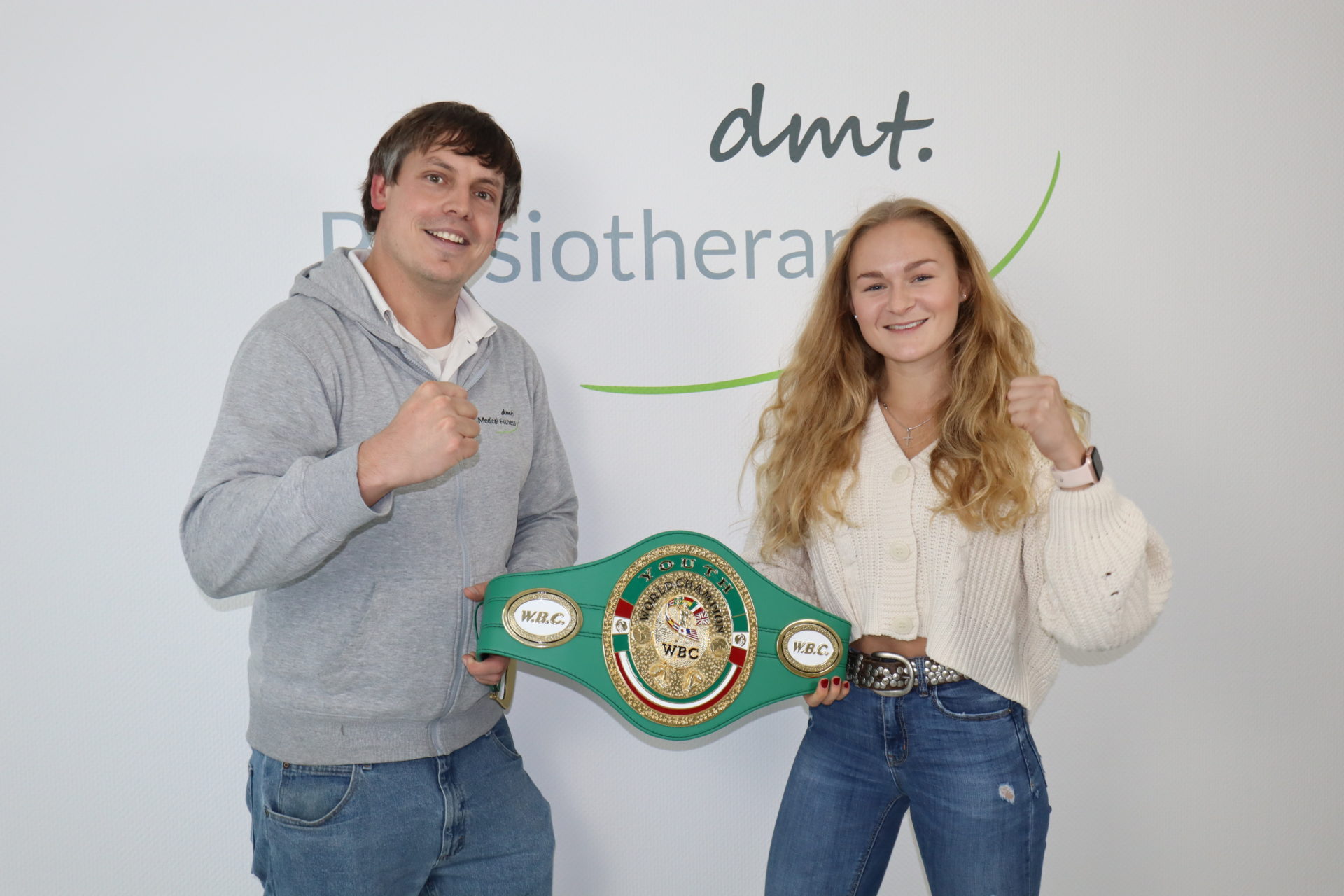Dennis und Sarah stehen vor dem dmt. Physiotherapie Logo und halten den Weltmeistergürtel.