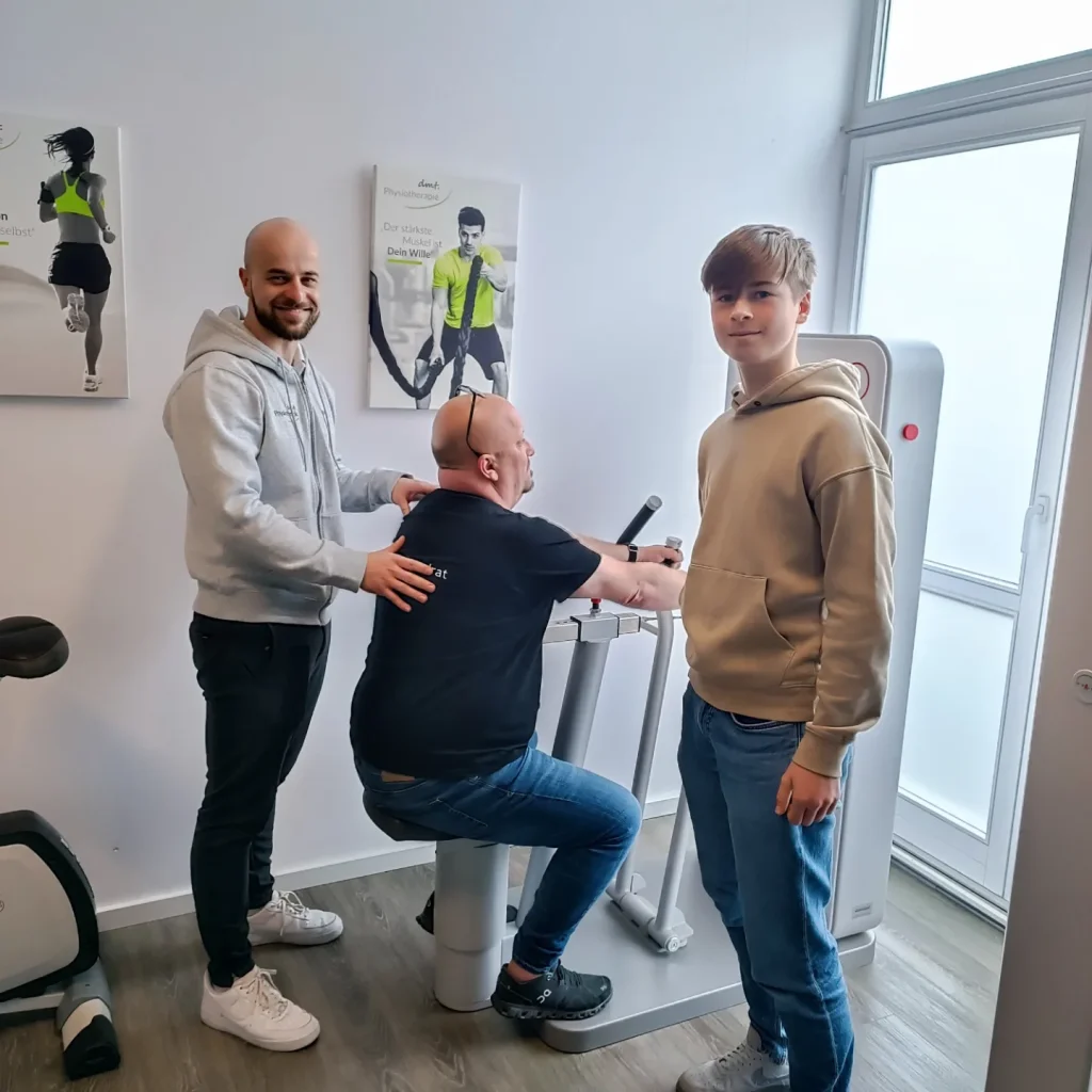Raimund leitet einen Patienten auf dem Rudergerät in der Physiotherapiepraxis an. Daneben steht ein Teilnehmer des Boy's Day.