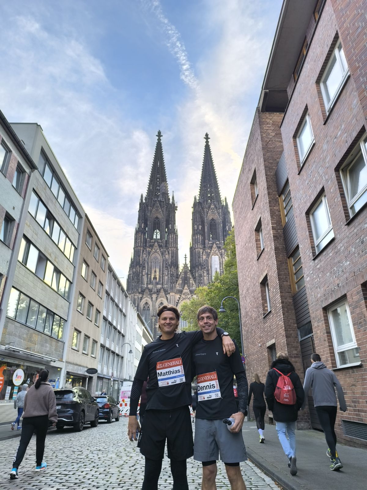 Links steht Matthias und rechts Dennis. Beide sind vor Ihrem Start beim Kölnmarathon mit dem Kölner Dom im Hintergrund zu sehen.