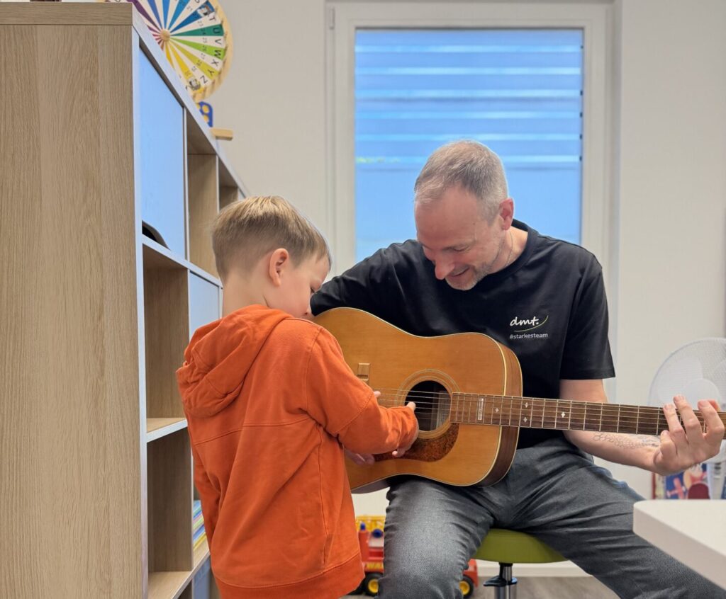 Logopäde Thomas aus Erftstadt hält eine Gitarre in der Hand. Ein Patient sthet vor ihm.