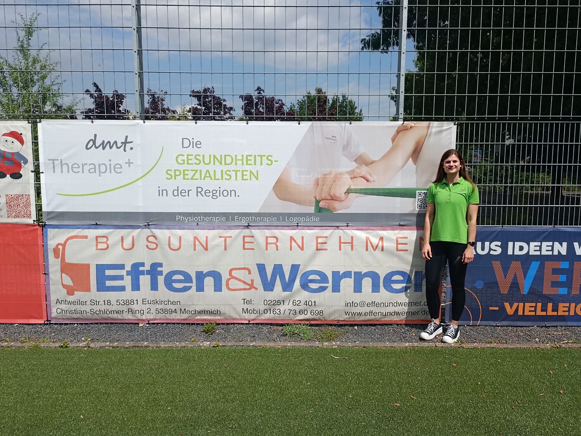 Katja steht rechts neben dem Banner der dmt. Therapie+ auf dem Sportplatz des JSG Erft 01 Euskirchen.
