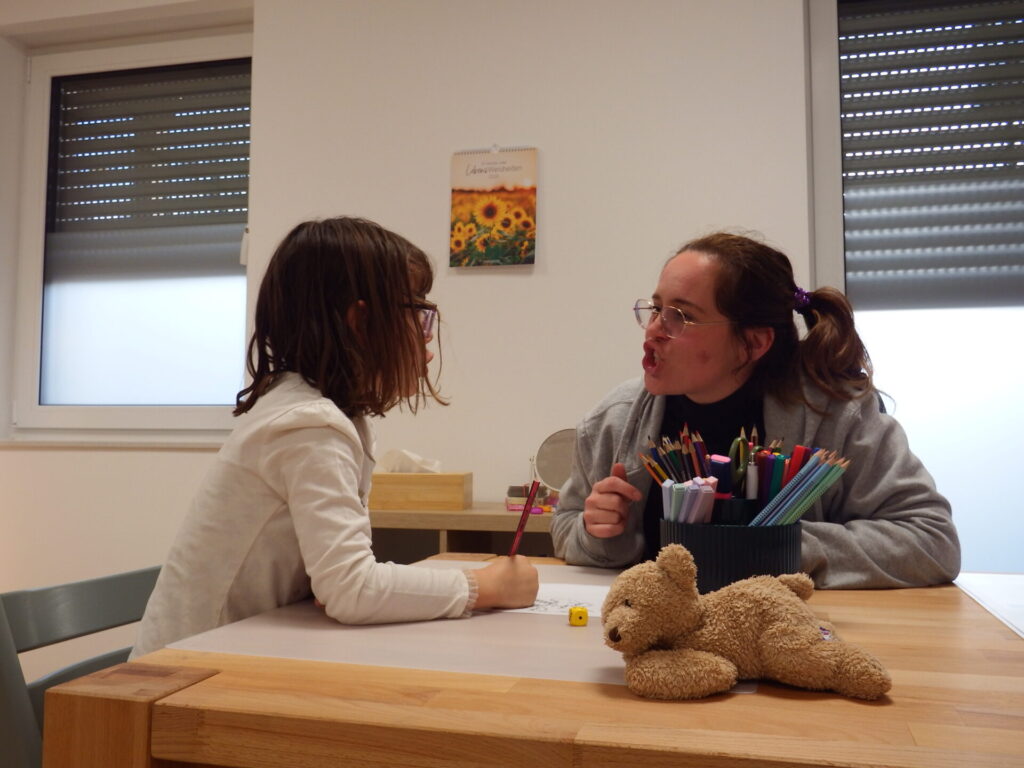 Jessi macht mit dem Mund Buchstaben nach. Die Patientin soll das nachahmen.