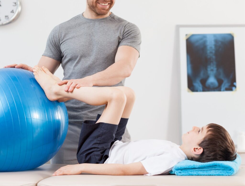 Kind liegt auf dem Rücken. Die Füße sind an einen Gymnastikball gestützt. Der Therapeut hält die Spunggelenke des Kindes sanft fest.