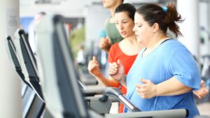 Eine übergewichtige Frau läft auf dem Laufband. Neben ihr steht ihre Trainerin uns spornt sie an.